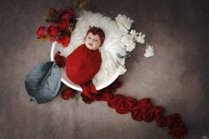 A baby is swaddled in red and surrounded by flowers.