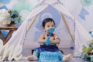 A baby boy sitting in front of a teepee with blue cake.