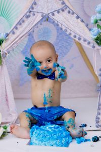 A baby is sitting in front of a teepee and smashing cake.