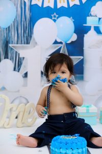 A baby boy eating cake in front of a blue backdrop.