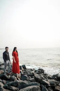 A couple standing on rocks near the ocean in red dress and black shirt.