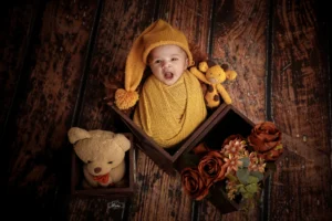 A baby in a yellow hat is sitting in a wooden box with teddy bears.