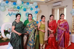 A group of women in saris and shootstanding in front of balloons in birthday party.