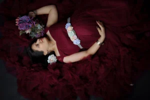A pregnant woman in a burgundy dress laying and posing with flowers.